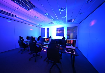 A modern gaming setup with multiple high-backed gaming chairs and monitors arranged in a room with a cool blue lighting. The computers have illuminated keyboards and glowing screens, creating a high-tech ambiance.