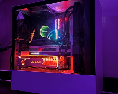 A gaming PC with a transparent side panel reveals internal components illuminated by RGB lighting. Key components visible include a graphics card, cooling system, and motherboard, all surrounded by multicolored LED lights. The case has a minimalistic design, set against a dark background that highlights the vibrant internal lighting.