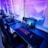 A row of modern gaming setups is arranged neatly along a window-lined wall. Each station includes a high-resolution monitor displaying vivid graphics, a mechanical keyboard with colored lighting, a gaming mouse, and an ergonomic chair. The room is illuminated with ambient blue and purple lights, creating a sleek and futuristic atmosphere.
