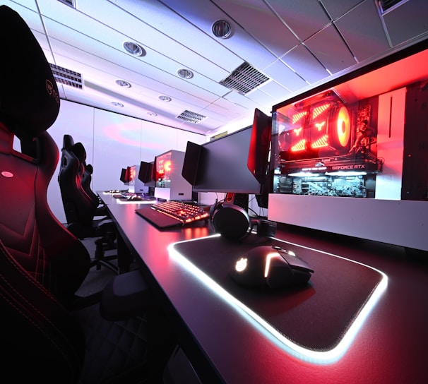 A row of high-end gaming computers on sleek desks, featuring RGB lighting and gaming accessories. The computers are housed in transparent cases with visible components, emitting a red glow. Comfortable gaming chairs with ergonomic design are positioned next to the desks, and the room's ceiling is modern with recessed lighting.