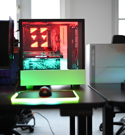 A modern gaming setup is displayed on a desk with multiple computer monitors and an open gaming PC tower. The tower is illuminated with red and green LED lights, showcasing a 'GeForce RTX' graphics card. A gaming mouse rests on a mousepad with glowing edges, and there is a keyboard with vibrant backlighting. The room appears well-lit with visible windows in the background.